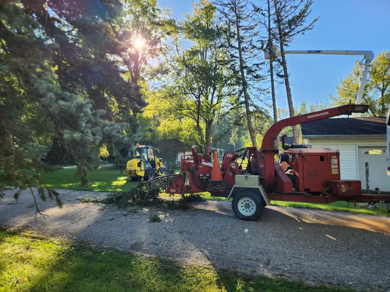 Red chipper parked on a gravel drive with branches being fed into it and a yellow loader behind